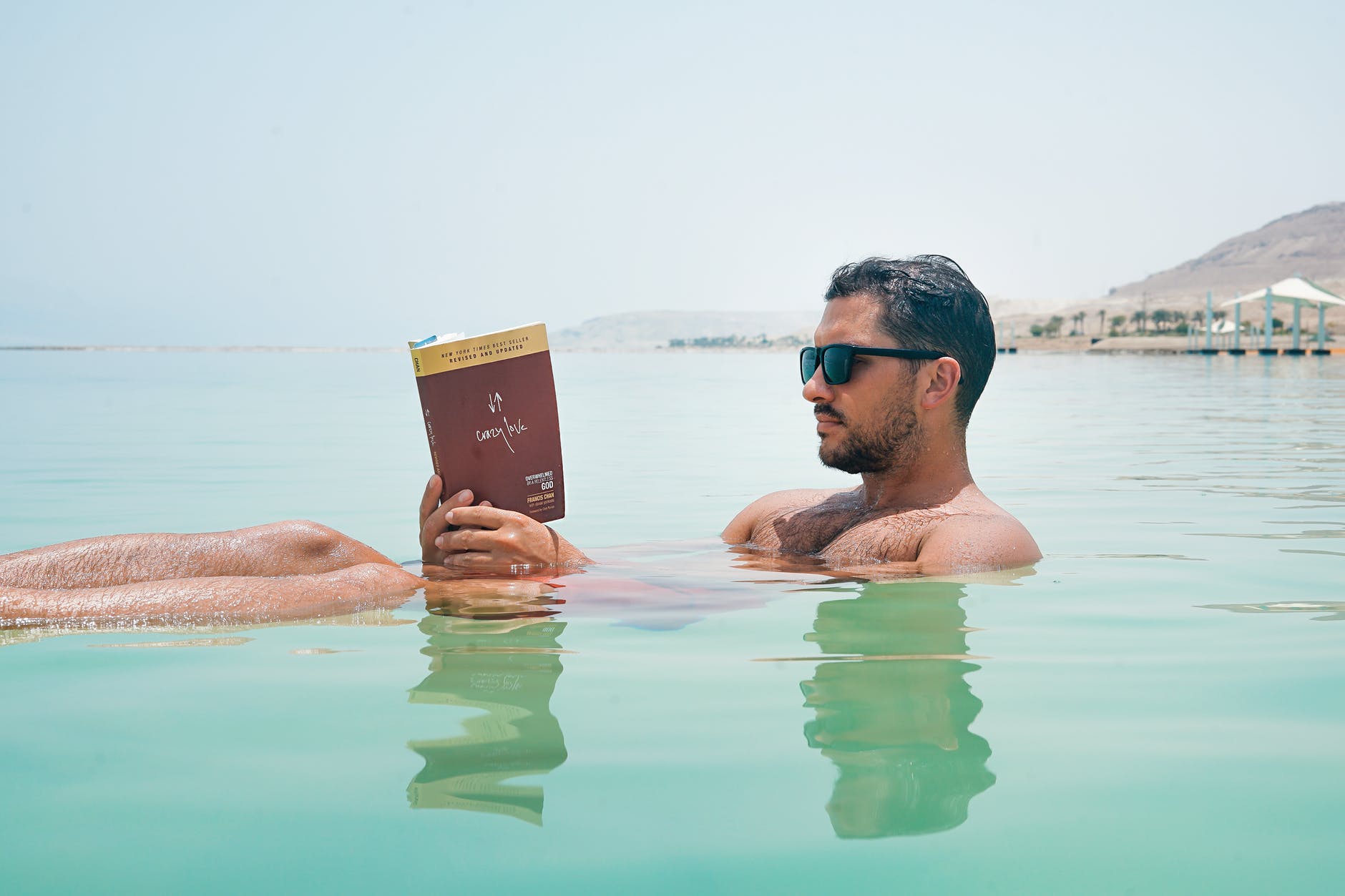 man wearing sunglasses reading book on body of water